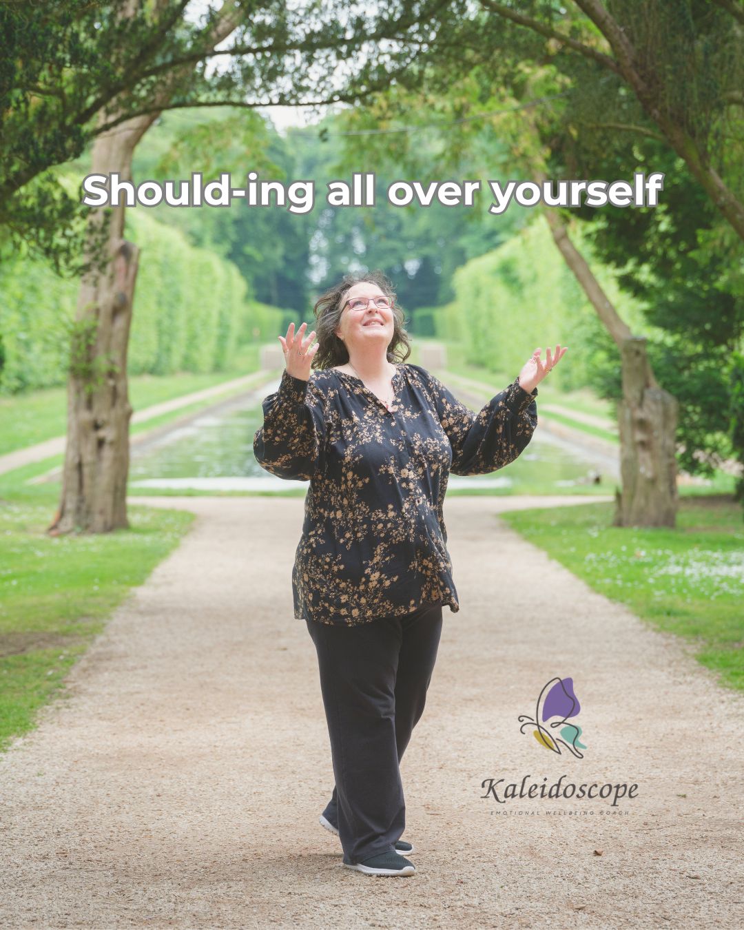 Photograph of Valerie Crozier-Nicholl standing on a tree-lined pathway with her hands raised and a thoughtful, slightly playful expression. The image features the title "Shoulding all over yourself" and accompanies a blog about self-compassion, self-criticism and noticing the language we use with ourselves.